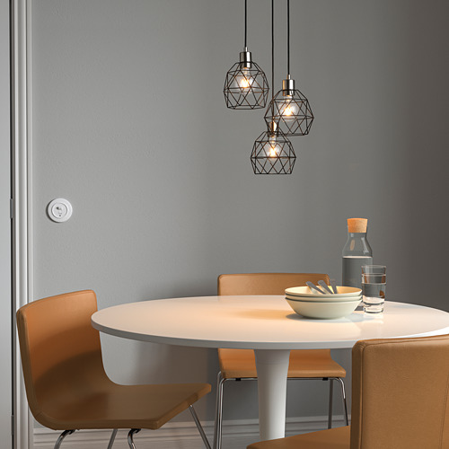 Moderno comedor con tres lámparas colgantes BRUNSTA, pequeñas pantallas geométricas de metal negro, sobre una mesa redonda de madera clara con sillas naranjas.