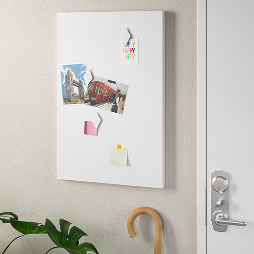 Tablero de notas magnético blanco con fotos y notas en una pared, junto a una puerta blanca con una manija plateada y un gancho de paraguas de madera que sostiene una planta verde.
