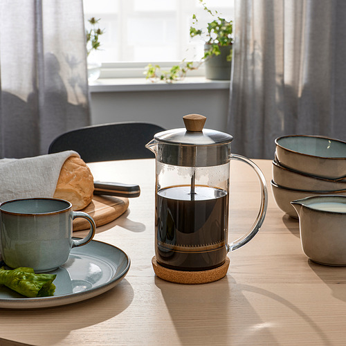 Mesa de cocina con prensa francesa, café, taza, pan y platos, ventana iluminada por el sol.