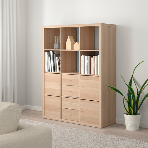 Estantería KALLAX con libros, decoración y planta. Sofá beige parcialmente visible.