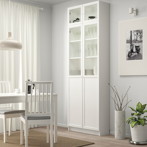 Estantería blanca BILLY en esquina, puertas de vidrio revelan platos, se eleva sobre la mesa de comedor, se adapta a una decoración minimalista.