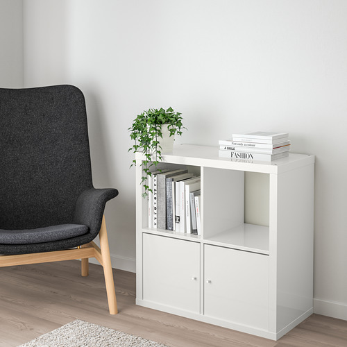 Estantería blanca KALLAX de ikea con libros y planta, junto a una gran silla gris sobre suelo de madera clara.