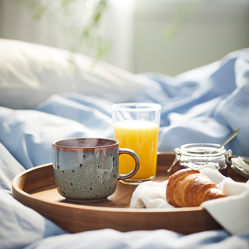 Bandeja de desayuno con taza GLADELIG, zumo de naranja, bollería, mermelada. La taza es azul, grande, de cerámica con diseño punteado.