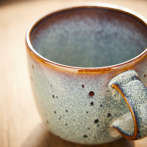 Taza artesanal con esmalte moteado y verdoso, paredes gruesas y mango resistente.