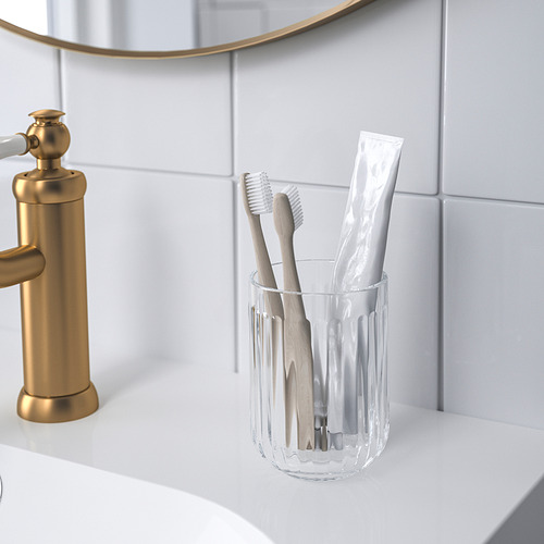 El soporte para cepillos de dientes de vidrio transparente, que sostiene cepillos de dientes y pasta de dientes, se encuentra cerca del grifo dorado.