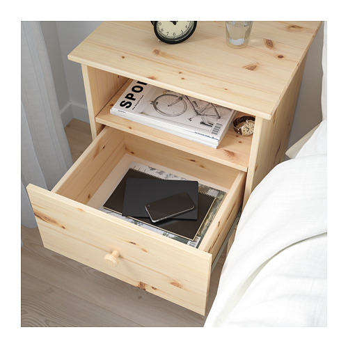 Mesita de noche de madera con cajón abierto que muestra una computadora portátil, un teléfono y una computadora portátil. El estante superior tiene una revista, un reloj y un vaso.