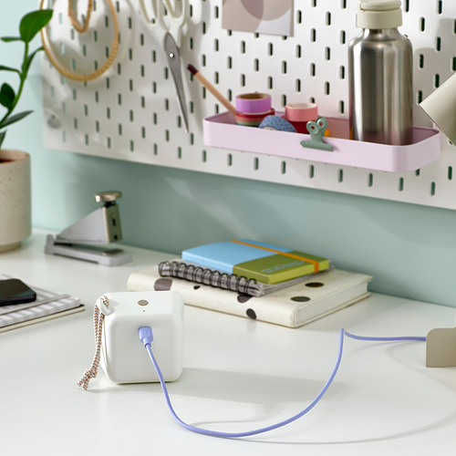 Un escritorio limpio con un cable usb-c blanco conectado a un pequeño dispositivo blanco, cerca de cuadernos y una planta.