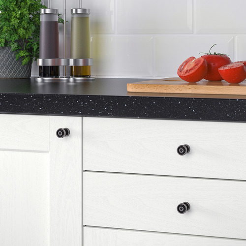 Dos tomates en una tabla de cortar, flanqueados por botellas con dispensadores, colocados sobre una encimera moteada de negro contra un salpicadero de azulejos blancos.