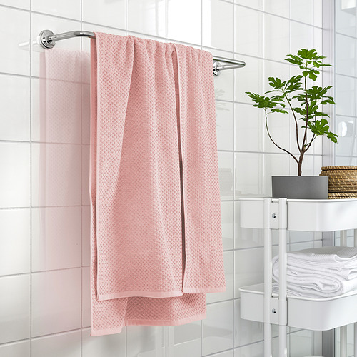 Baño moderno con toallas de color rosa claro en barra, toallas blancas cuidadosamente dobladas y una pequeña planta en maceta en un estante blanco contra la pared de azulejos blancos.