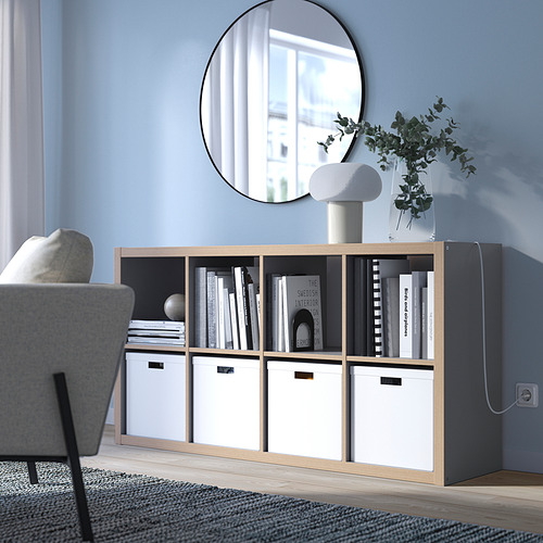 Estantería KALLAX en color gris claro con madera clara, rectangular, diseño minimalista. Se sienta bajo, cabe debajo de un espejo redondo, sosteniendo libros y una lámpara.