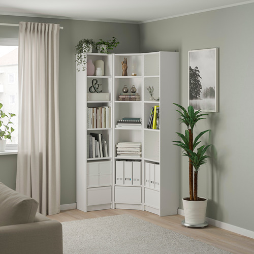 Estantería esquinera blanca BILLY llena de libros y decoración, colocada cerca de un gran ventanal con cortinas beige, y acompañada de una alta planta de interior.