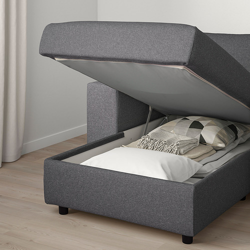 Un sofá tapizado gris con espacio de almacenamiento debajo, parcialmente abierto para revelar mantas y almohadas en el interior.