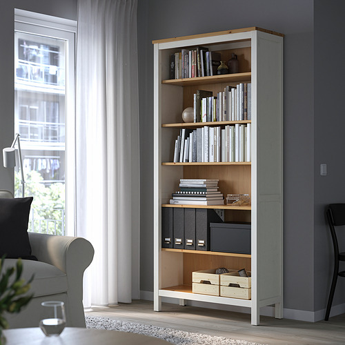 Estantería alta y delgada HEMNES en blanco/pino, llena de libros y decoración, junto a una ventana y un sofá gris.