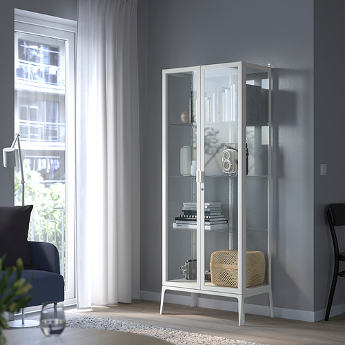 Armario con puerta de vidrio blanco con libros y decoración, cerca de la silla azul, habitación tranquila.