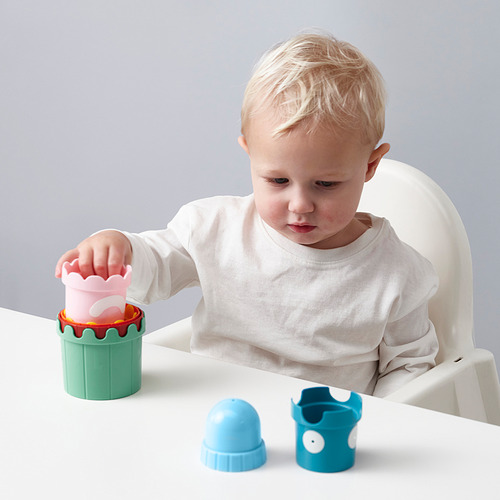 Un niño pequeño se sienta en una silla alta jugando con tazas apilables de colores, explorando formas y colores.