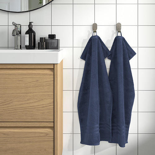 Baño ordenado con tocador de madera. Las grandes toallas BROKGLIM azul marino cuelgan cuidadosamente en los ganchos de la pared. Los azulejos blancos contrastan con los cálidos armarios de madera.