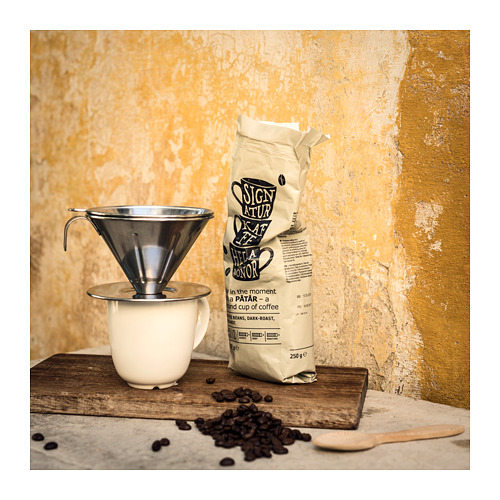 Una escena de café con un filtro plateado reutilizable, una taza blanca y frijoles marrones. La bolsa lee disfruta del momento con un fondo amarillo.