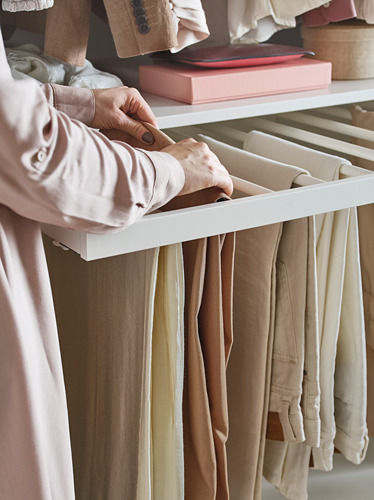 Una persona organiza pantalones beige con la percha de pantalón KOMPLEMENT, blanca, de acero y tablero de fibra, en un armario.