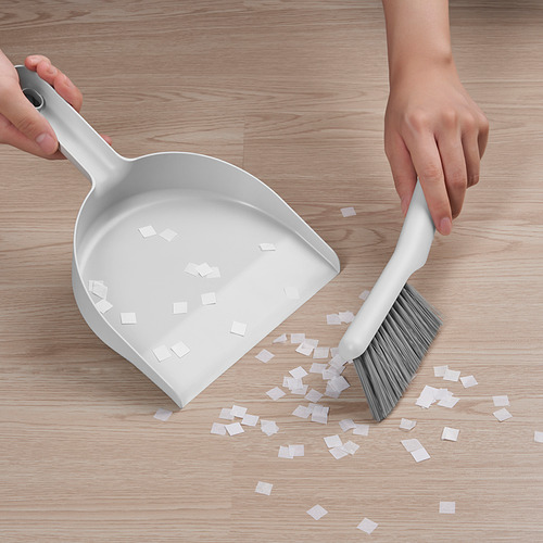 La persona limpia el piso con un recogedor y un cepillo de PEPPRIG blanco, recogiendo pequeños trozos de papel; Muestra el diseño de ángulos efectivo para la limpieza de esquinas.