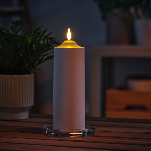 Una sola vela alta y blanca SMÅLOM sobre una mesa de madera, encendida, rodeada de planta e interior borrosos.