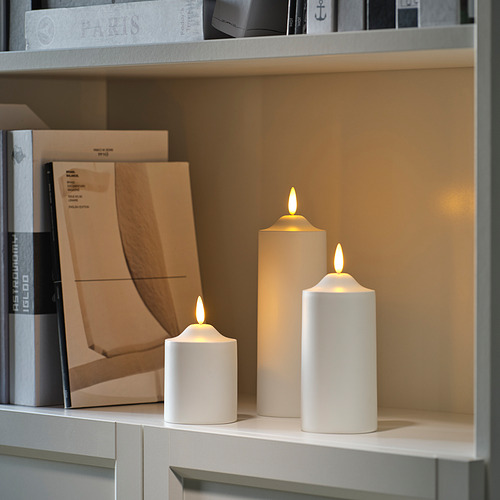 Tres velas SMÅLOM led en beige claro sobre una estantería, de diferentes tamaños, iluminadas, rodeadas de libros.