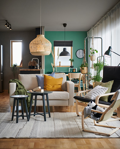 Acogedora sala de estar con sofá beige, almohadas de colores, piso de madera y pared verde. Alfombra brillante, lámpara colgante de mimbre y plantas colgantes.