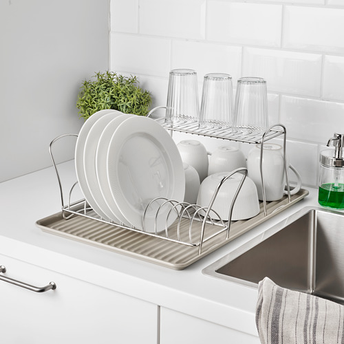 Un estante para platos cromado contiene platos, tazas y vasos blancos, colocados sobre un fregadero para que se sequen al aire. Se adapta a un escurridor para encimeras limpias.