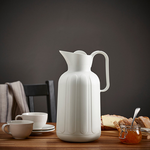 Frasco blanco sobre mesa de madera con taza de té, tazas, platillos, mermelada, pan y tabla de cortar.