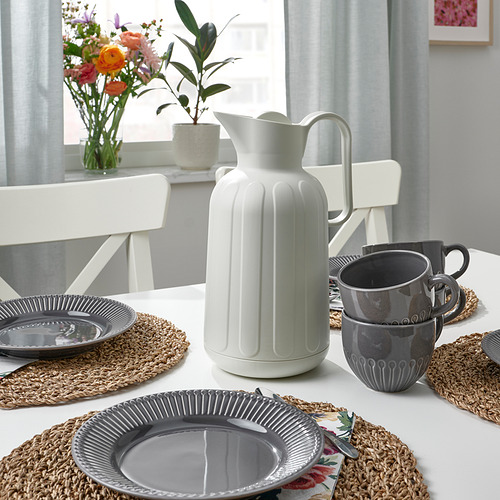 Mesa con platos y tazas grises, jarra blanca, manteles individuales de pastos marinos, flores y plantas en macetas.