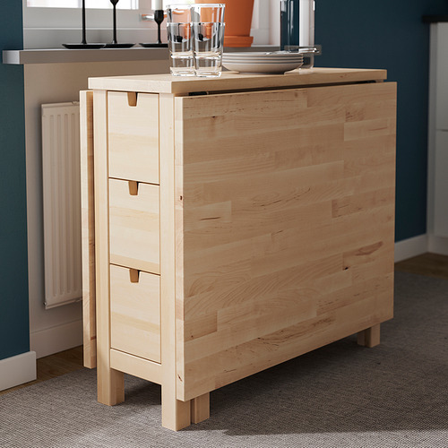 Una mesa de madera clara con tres cajones y dos vasos de cristal encima, está apoyada contra la pared azul.