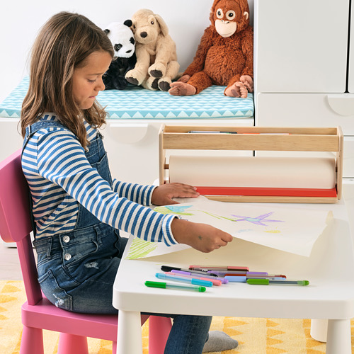Un niño se sienta en una mesa dibujando con rotuladores de colores, utilizando el rollo de papel MÅLA de un caballete. Los animales de peluche se sientan cerca.