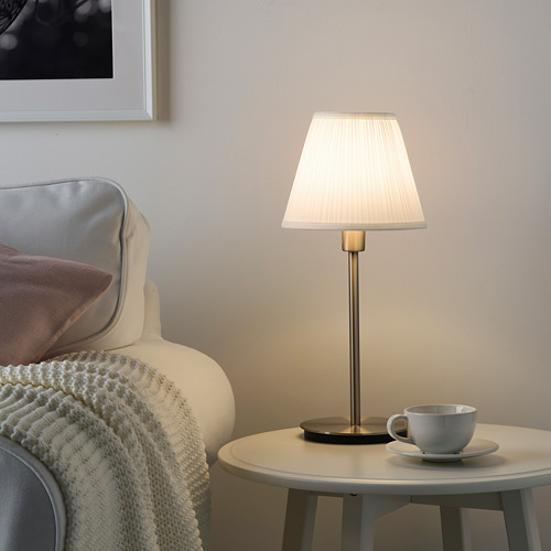 Pequeña y moderna lámpara SKAFTET con base dorada, pantalla redonda blanca en la mesa auxiliar junto a una silla acogedora.