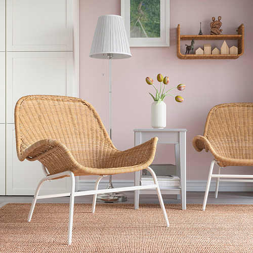 Dos sillones de mimbre, patas blancas, alfombra, mesa auxiliar con flores, lámpara, estantería con decoración.