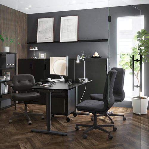 Modernos escritorios negros de oficina con sillas ergonómicas, rodeados de detalles en madera oscura y plantas decorativas. Dos mapas de la ciudad enmarcados adornan la pared.