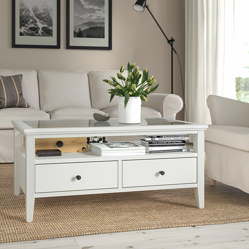 Mesa de centro IDANÄS blanca con tapa de cristal, cajones y flores. Centrado en una sala de estar con un sofá beige y alfombra.