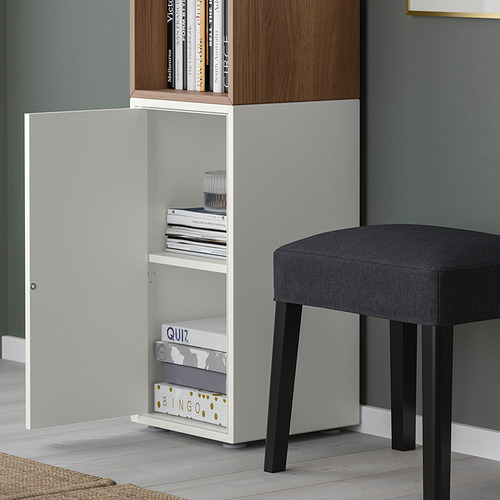 Mueble de almacenamiento EKET blanco con puerta abierta, juegos y libros; Taburete tapizado gris al lado.