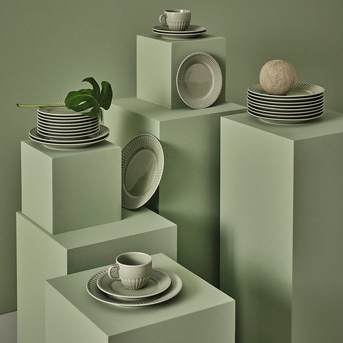 Exhibición de vajilla moderna, que incluye tazas, platillos, platos STRIMMIG en verde salvia. Cerámica con diseños limpios y artesanales, dispuestos en varias plataformas.