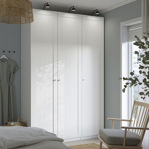 Modern white PAX wardrobe in bedroom, tall with sleek lines. Wood chair, clothes, and big window beside it.