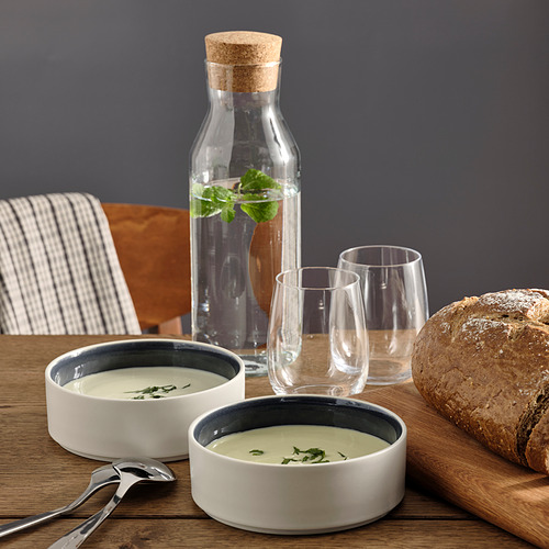 Dos tazones de sopa de cerámica, dos vasos, una jarra de vidrio con agua con hierbas, silla de madera, servilleta a rayas, barra de pan sobre una mesa de madera.