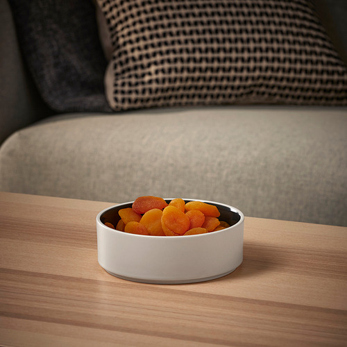 Un pequeño cuenco de cerámica blanca lleno de frutos secos de color naranja y amarillo se encuentra sobre una mesa de madera clara.