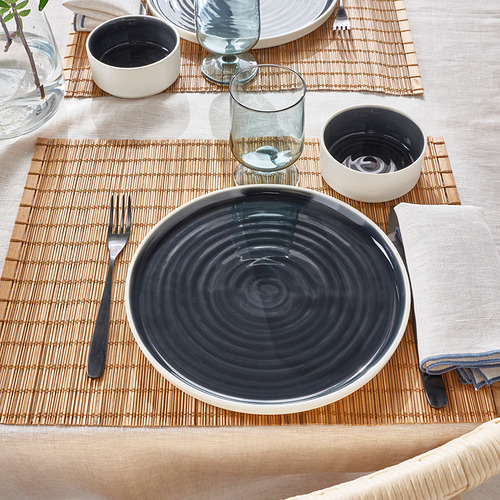 Mesa con platos minimalistas OMBONAD. Incluye un plato de cerámica, tazones pequeños, vidrio, tenedor y servilleta sobre un tapete tejido.