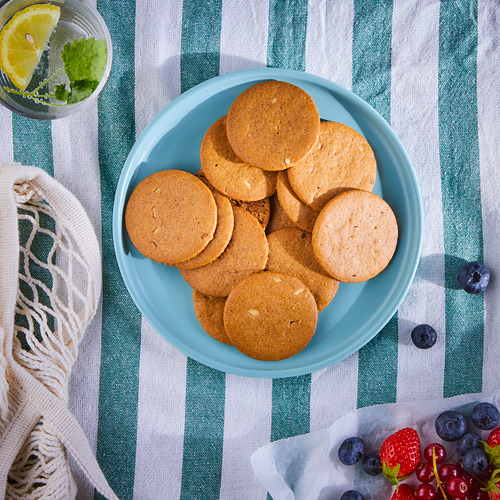 Un plato azul de galletas de jengibre marrón se encuentra sobre un paño a rayas verdes, junto a una bebida con limón y menta, y una canasta de bayas mixtas.