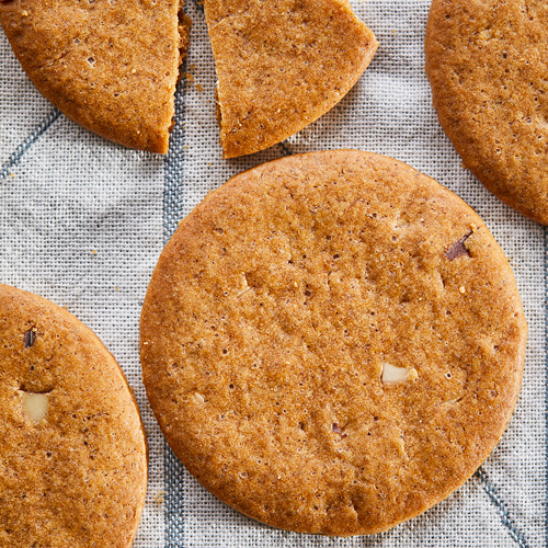 Seis galletas de jengibre KAFFEREP de color marrón dorado, redondas, crujientes y moteadas con diminutas migas de almendra, reposando sobre un paño gris claro.