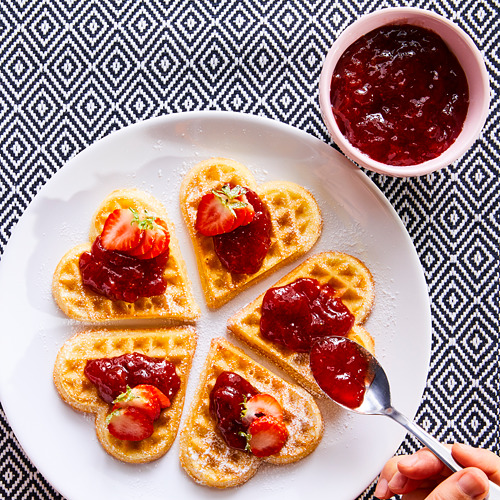 La imagen muestra gofres en forma de corazón con mermelada roja y fresas en un plato blanco con una cucharada de mermelada, sobre un mantel estampado en blanco y negro.
