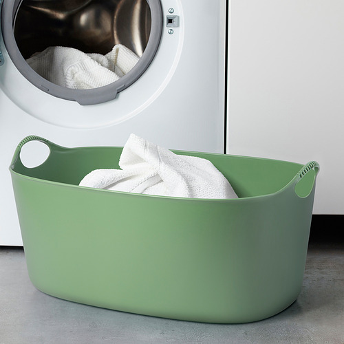 Cesta de lavandería flexible verde con toallas blancas cerca de una lavadora con puerta abierta.