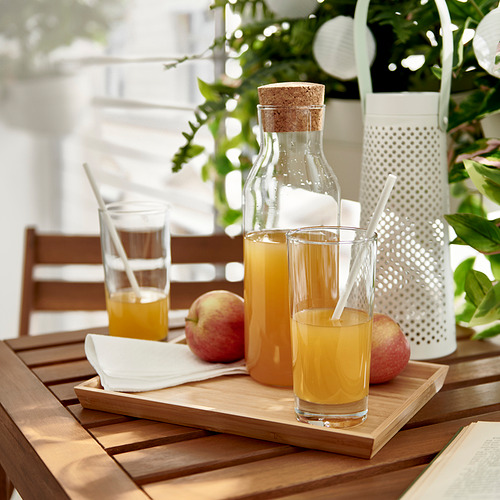 Dos vasos de jugo de naranja en una bandeja de madera con manzanas, servilletas y pajitas. El fondo tiene una linterna y plantas verdes.