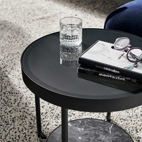 Mesa auxiliar circular negra con tapa de cristal, patas metálicas y estante efecto mármol. Un vaso de vidrio, un libro y vasos colocados sobre él.