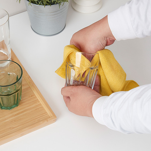 Persona limpiando vasos con un paño amarillo sobre una mesa blanca. Dos vasos, planta y tabla de madera cerca.