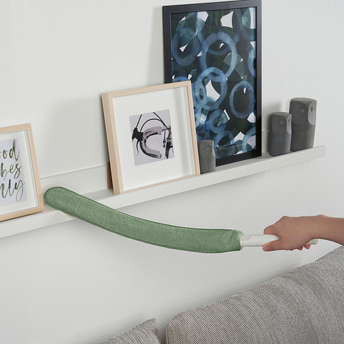 Una mano que sostiene un recambio de plumero de microfibra PEPPRIG verde, estirándolo horizontalmente sobre un estante blanco con decoración de pared.