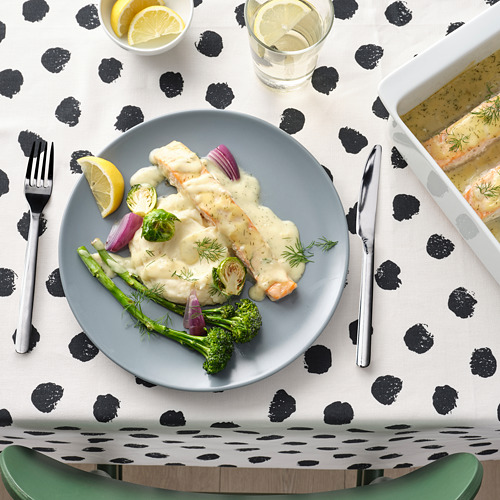 Una comida de salmón, verduras y salsa en un plato con cubiertos sobre un mantel de lunares blancos y negros.